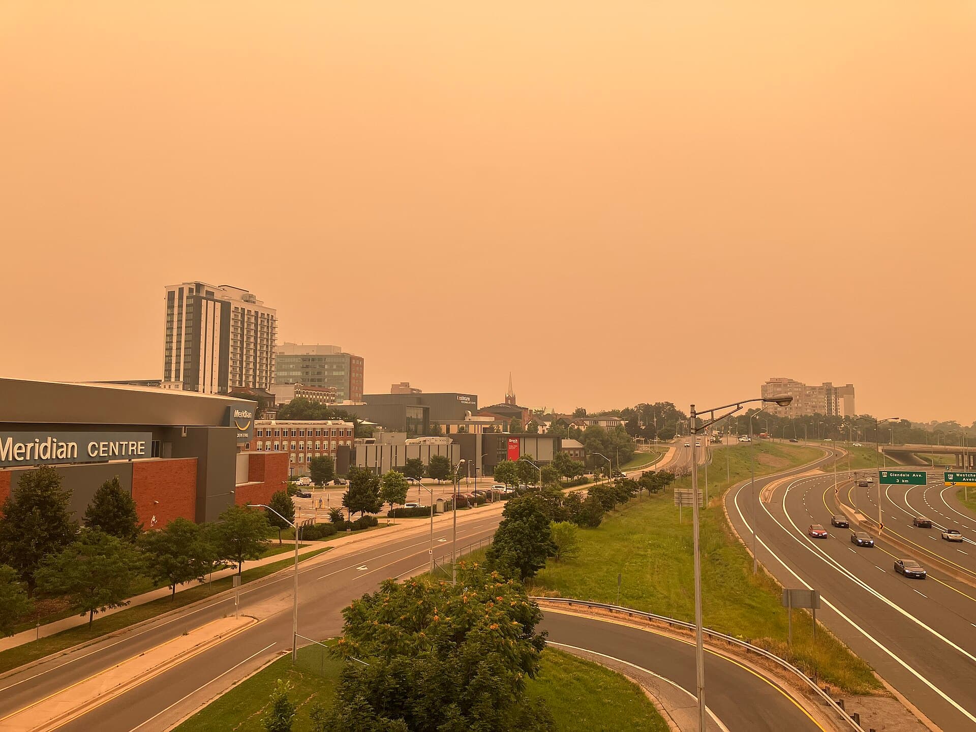 The city of St. Catharines, Ontario under a dark orange sky of wildfire smoke, cars driving with headlights on in the middle of the day